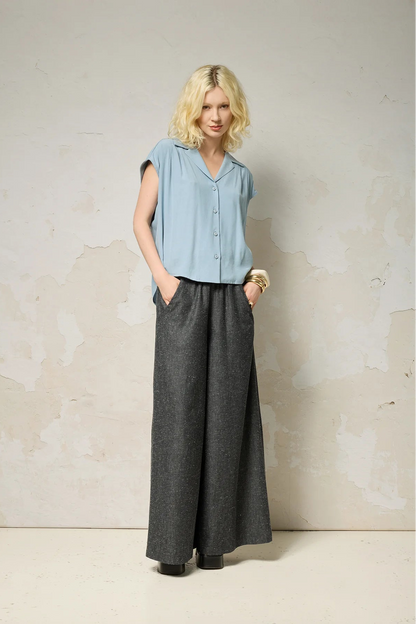 A woman wearing the Hemera Shirt by Cokluch in Sky, with a V-neck collar, button front, short extended sleeves, and a relaxed fit. The hem is hip-length in the front and longer at the back. She's wearing it with denim pants and standing in front of a grey background. 