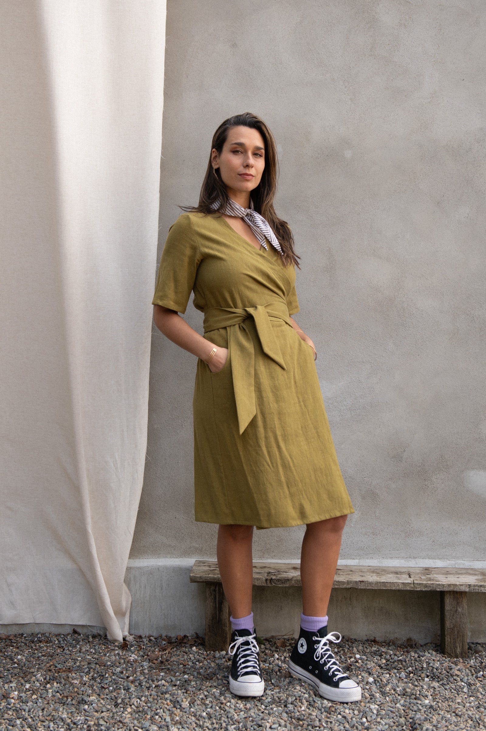 A woman wearing the Helleborus wrap Dress by Kazak in Mousse, standing outside by a white curtain and a cement wall. 