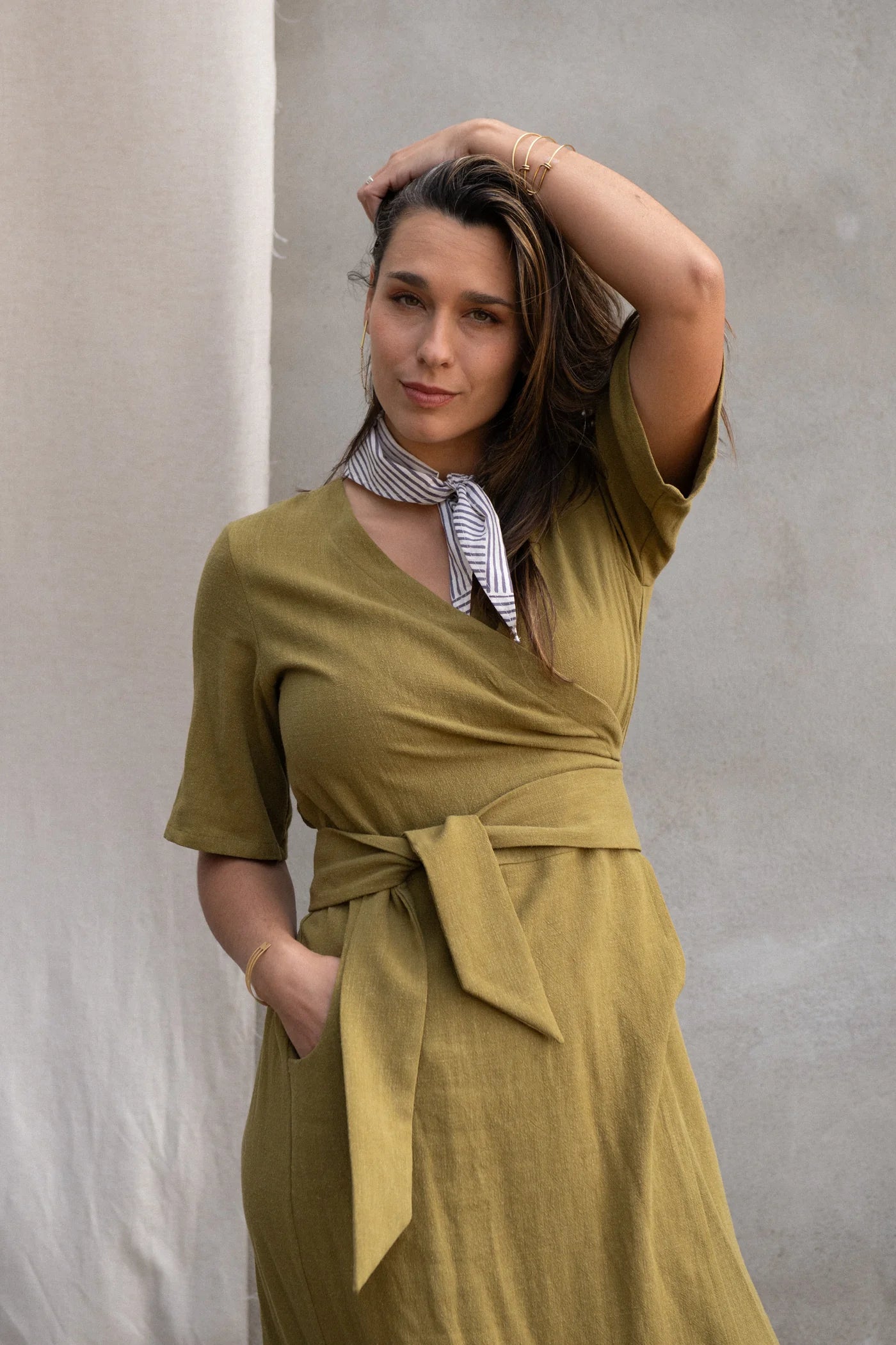 A woman wearing the Helleborus wrap Dress by Kazak in Mousse, standing outside by a white curtain and a cement wall. 