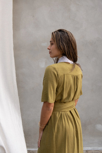 Back view of a woman wearing the Helleborus wrap Dress by Kazak in Mousse, standing outside by a white curtain and a cement wall. 