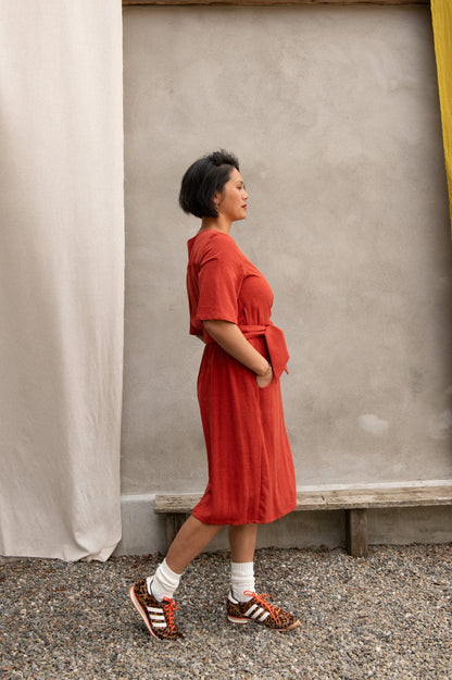 Side view of a woman wearing the Helleborus wrap Dress by Kazak in Paprika, standing outside by a white curtain and a cement wall. 