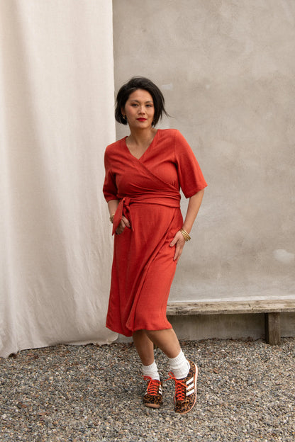 A woman wearing the Helleborus wrap Dress by Kazak in Paprika, standing outside by a white curtain and a cement wall. 