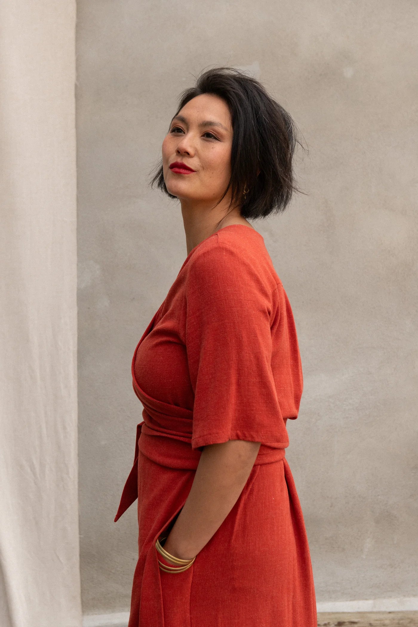 Side view of a woman wearing the Helleborus wrap Dress by Kazak in Paprika, standing outside by a white curtain and a cement wall. 