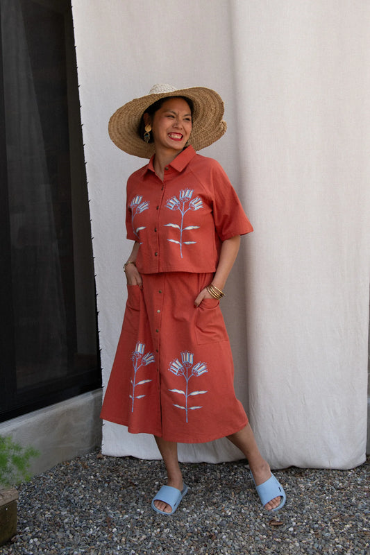 A woman wearing the Nymphea Screenprinted Skirt by Kazak in Red Pysanka with the matching Hosta top, she is standing outside in front of white curtain. 