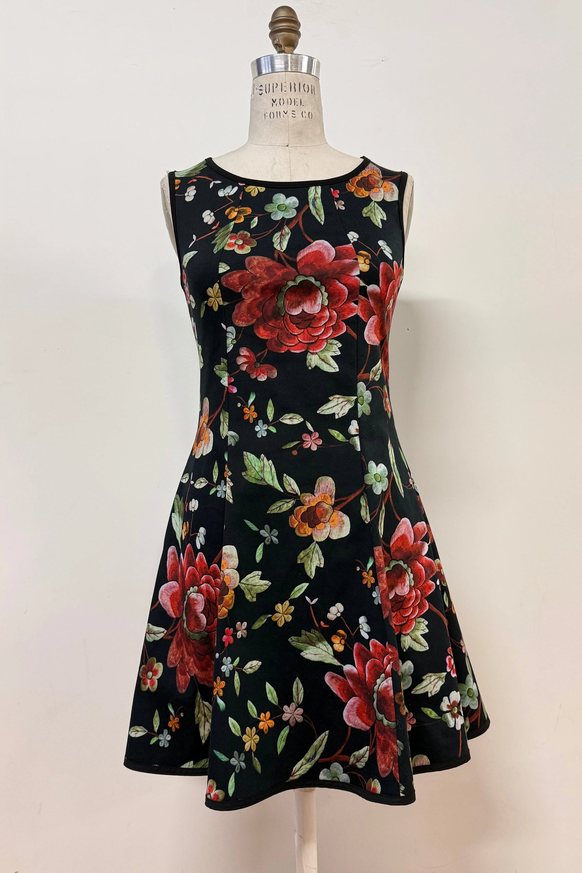 The Silba Dress FW26/27 by Kollontai in Black Floral, a short sleeveless dress with princess seams and an above the knee skirt, is shown on a mannequin against a white background. 