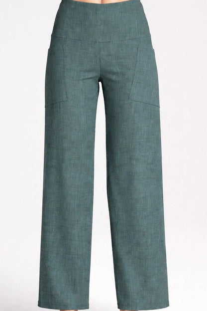 Waist down view of a woman wearing the Kollontai Susan Pants in Green, standing in front of a grey background. 