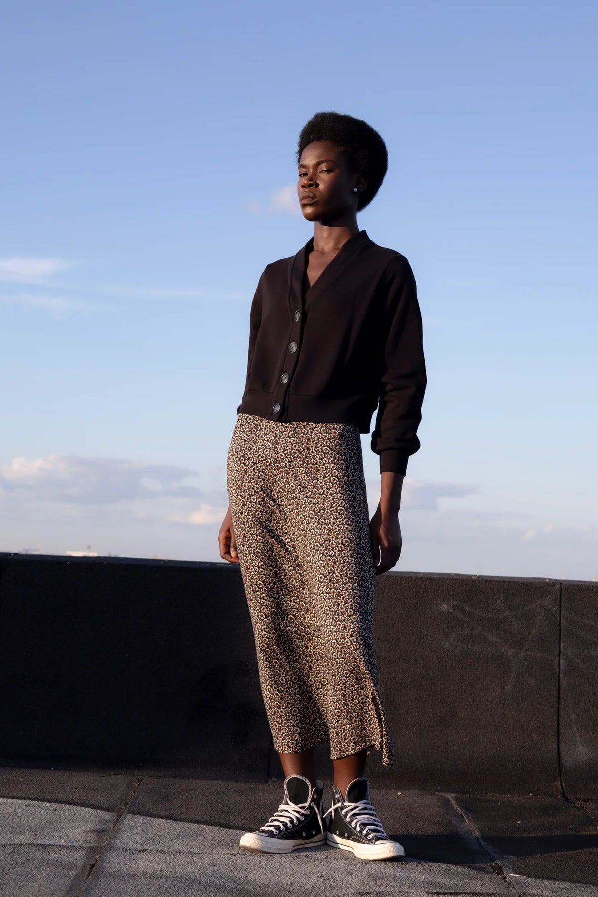 A woman wearing the Crystal Long Skirt by Eve Lavoie in Cherry Blossom print, an A-line maxi skirt. She's wearing it with a cardigan and sneakers and standing outdoors by a low concrete wall. 