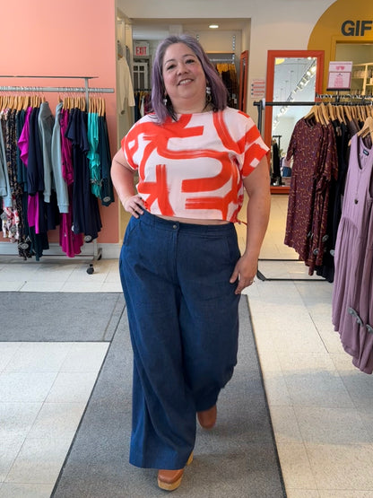 A woman wearing the Nyx top from Cockluch in Red Shodo, a short sleeved, loose fitting top with a drawstring detail at the bottom hem. She is wearing it with the atisse Trousers  by Melow and standing inside Flock. 