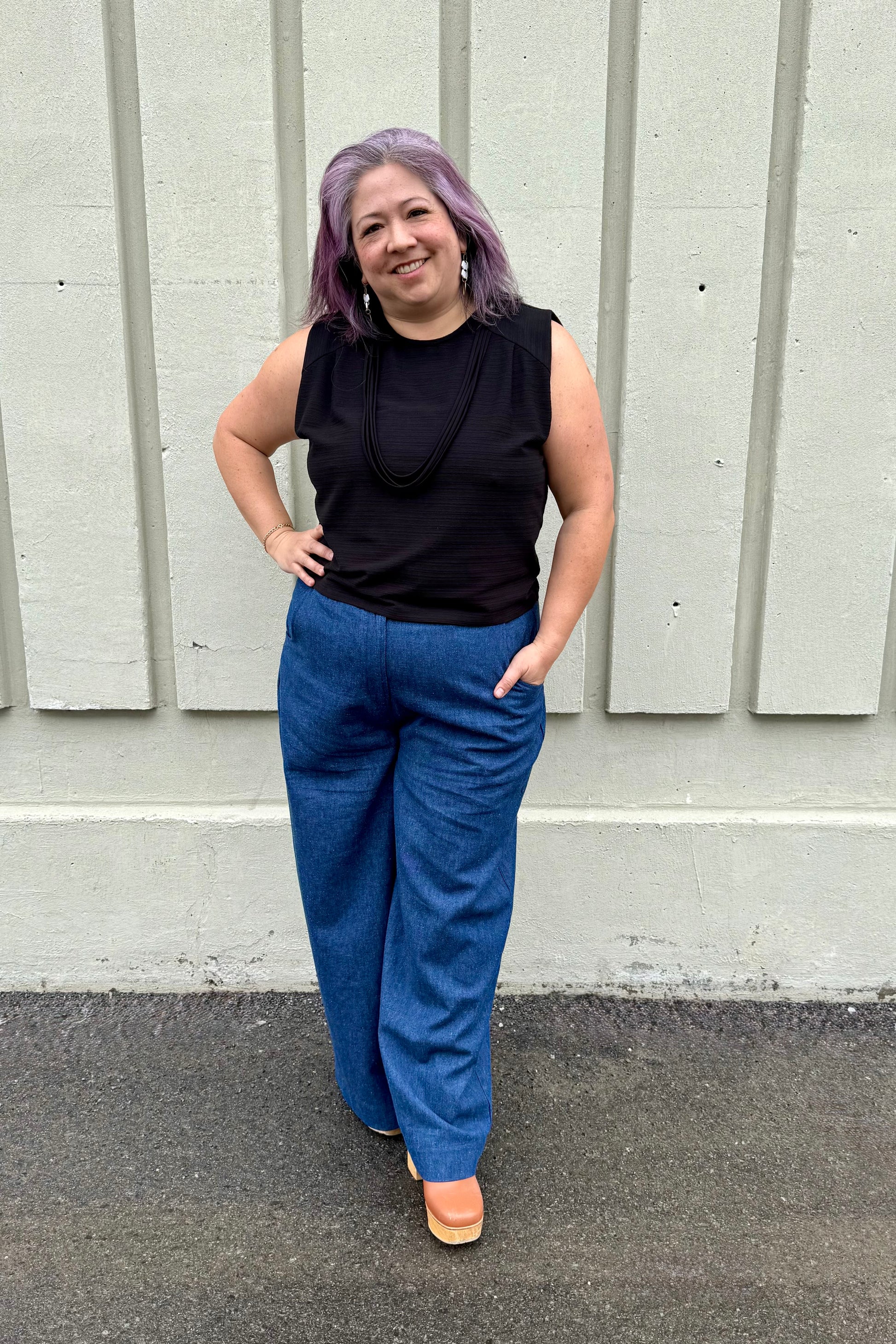 A woman wearing the Path Top from Cokluch in Black, a sleeveless top with line details at the shoulders and decorative straps that attach at each shoulder and hang to the front. She is wearing it with the Matisse pants by Melow and standing outside in front of a panelled wall. 