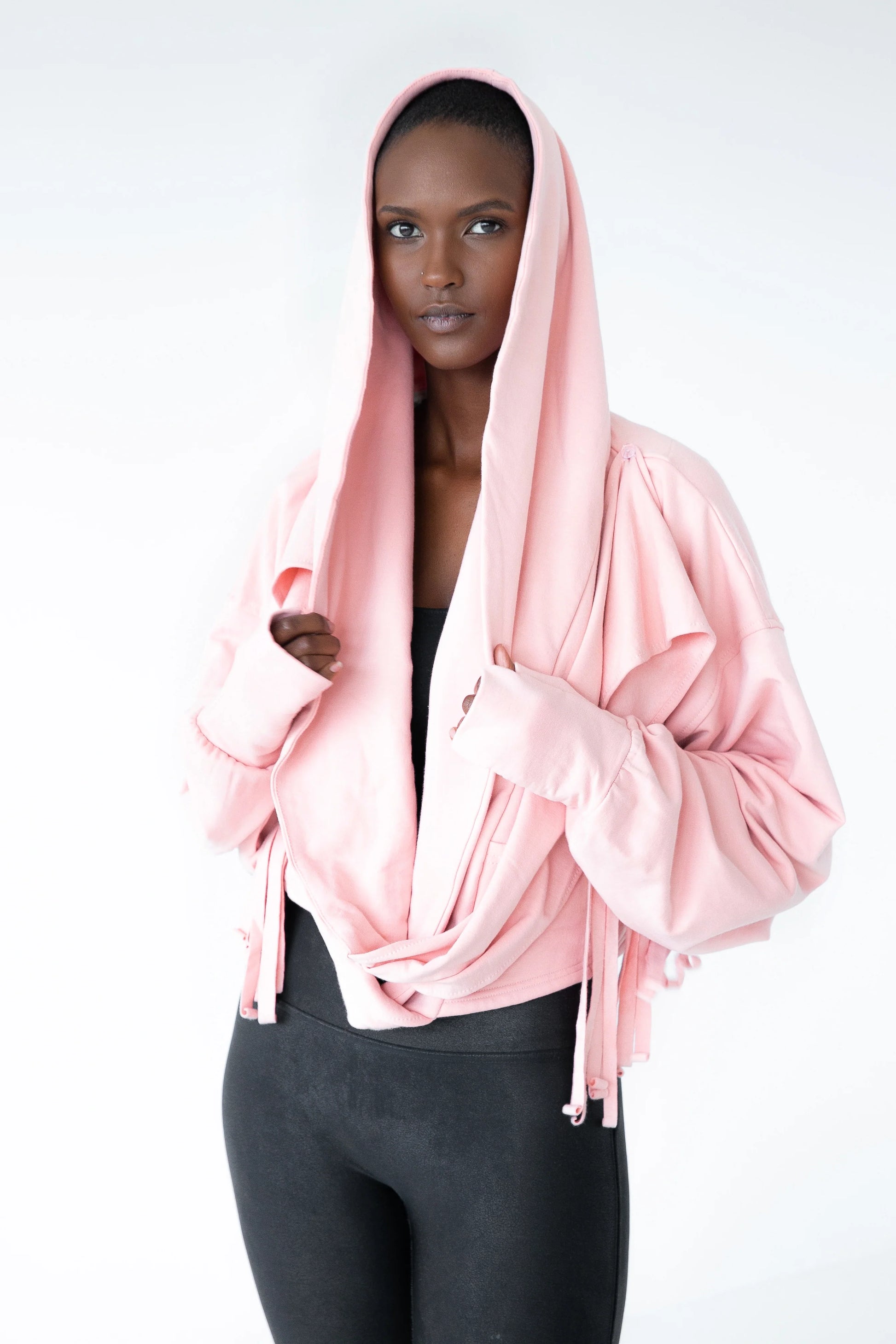A woman wearing the Heaven Bamboo/Cotton Cropped Wrap from Stacey Martin Lifestyle in Blush Pink, a cropped hoody with a fringed collar scarf that can be tied various ways. She is wearing it with black leggings and standing in a white room. 