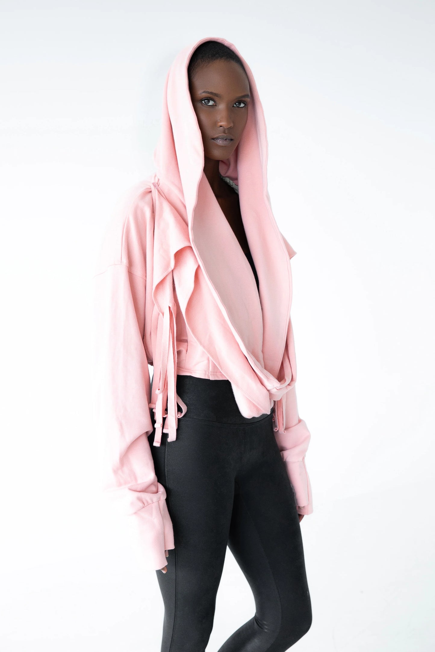 A woman wearing the Heaven Bamboo/Cotton Cropped Wrap from Stacey Martin Lifestyle in Blush Pink, a cropped hoody with a fringed collar scarf that can be tied various ways. She is wearing it with black leggings and standing in a white room. 