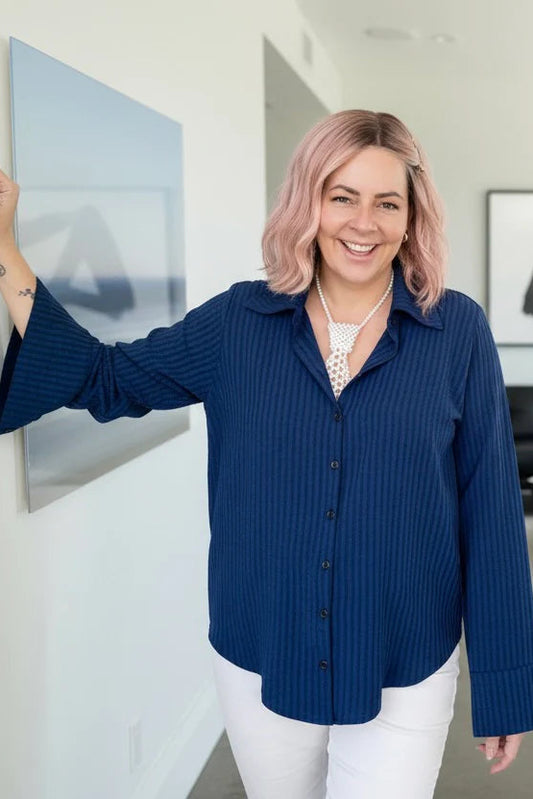 A woman wearing the Blue Striped Blouse by Dotty, a classic button front shirt with a collar and extra wide cuffs. She is wering it with white pants and standing in a room with art on the walls. 