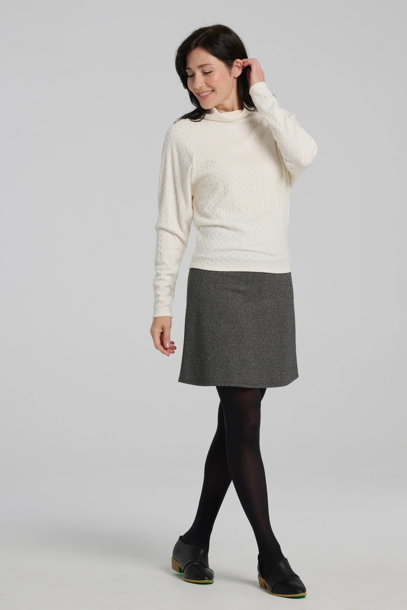 A woman wearing the Plume Sweater by Kollontai in White, a cable knit turtleneck with long tapered sleeves and a narrow hem for a bloused look. She is wearing it with a grey skirt and standing in front of a grey background. 