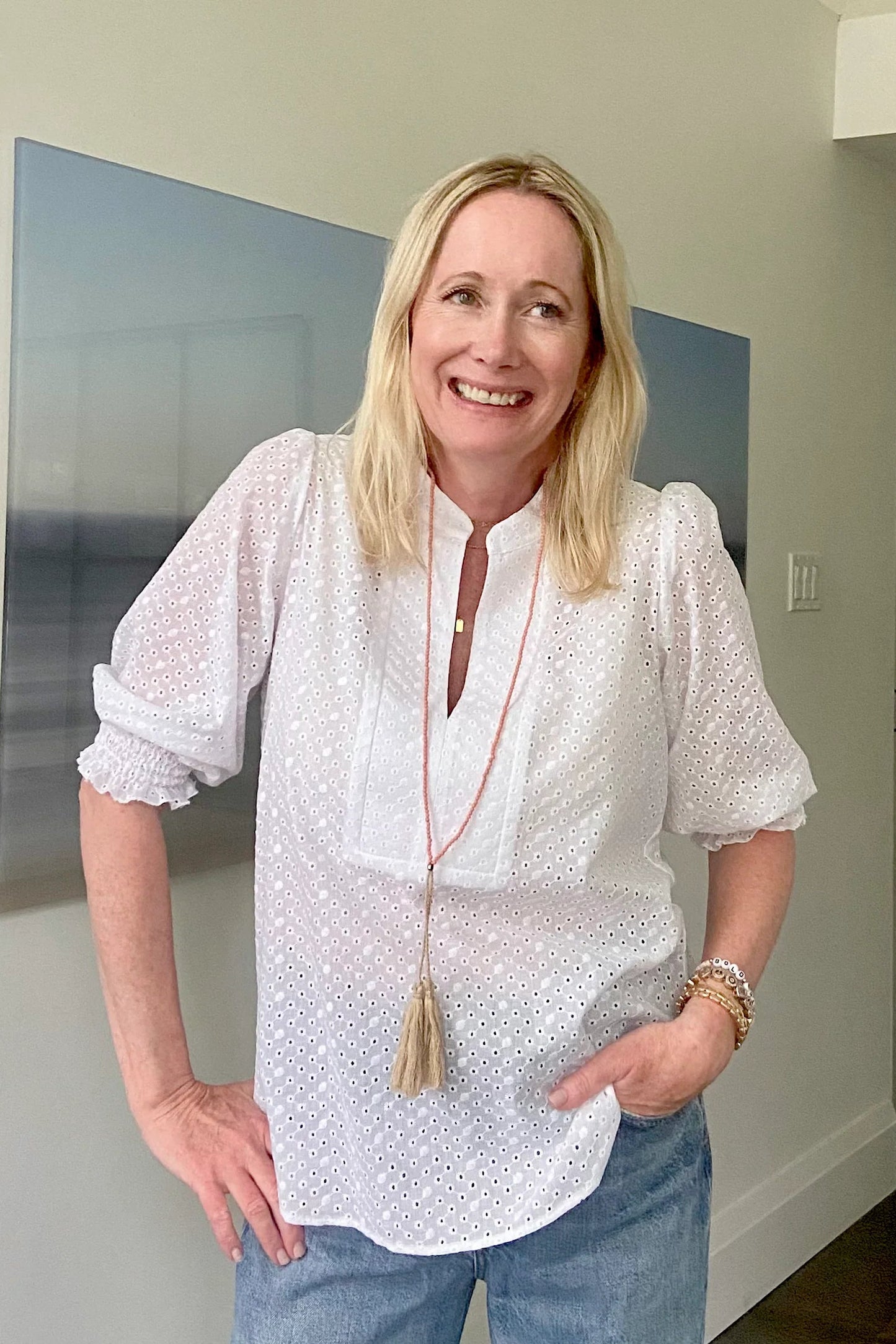A woman wearing the Eyelet Summer Blouse by Dotty in White, a pullover top with a banded v-neck and 3/4 smock sleeves. She is wearing it with jeans and standing indoors by a painting.