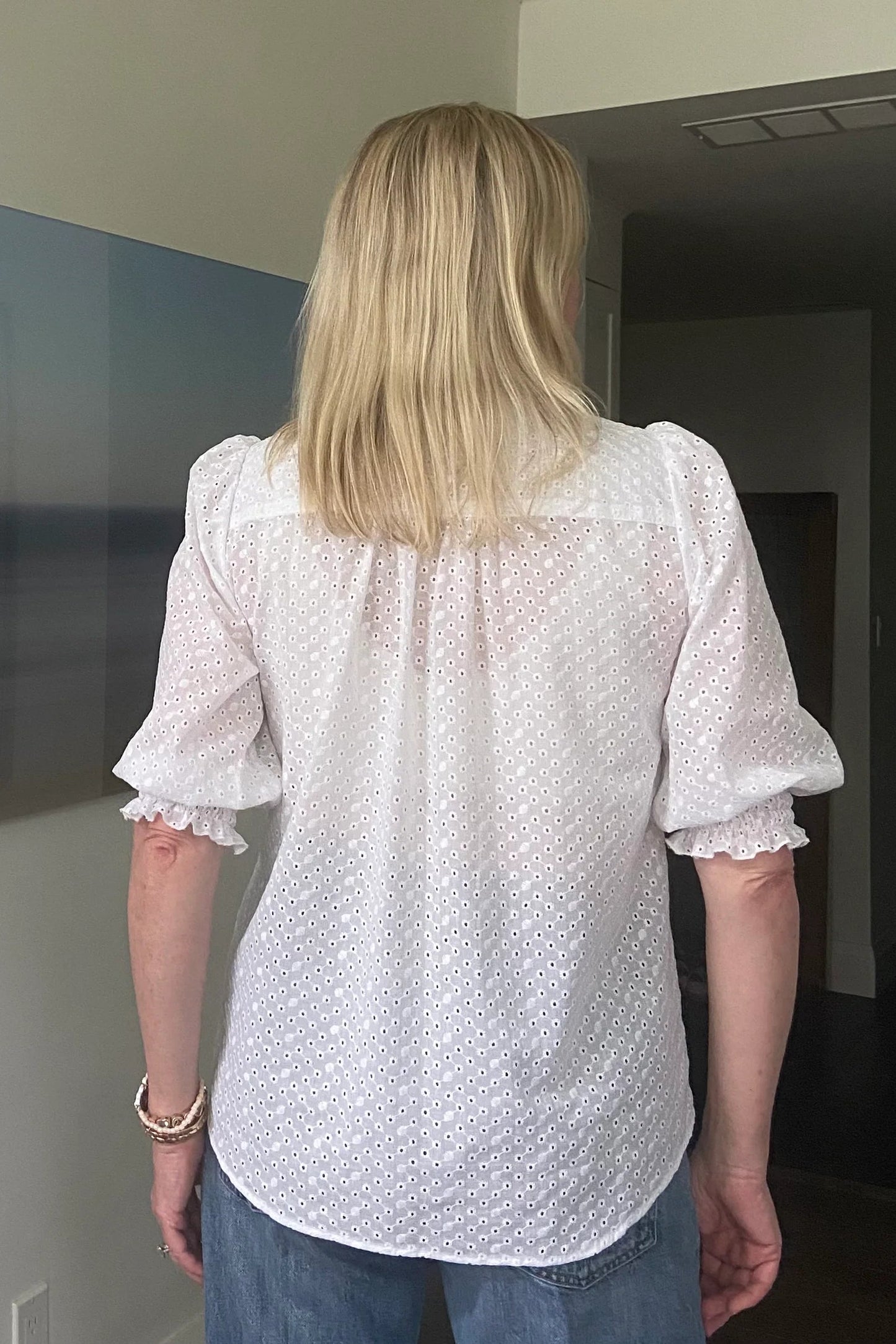 Back view of a woman wearing the Eyelet Summer Blouse by Dotty in White, a pullover top with a banded v-neck and 3/4 smock sleeves. She is wearing it with jeans and standing indoors by a painting.