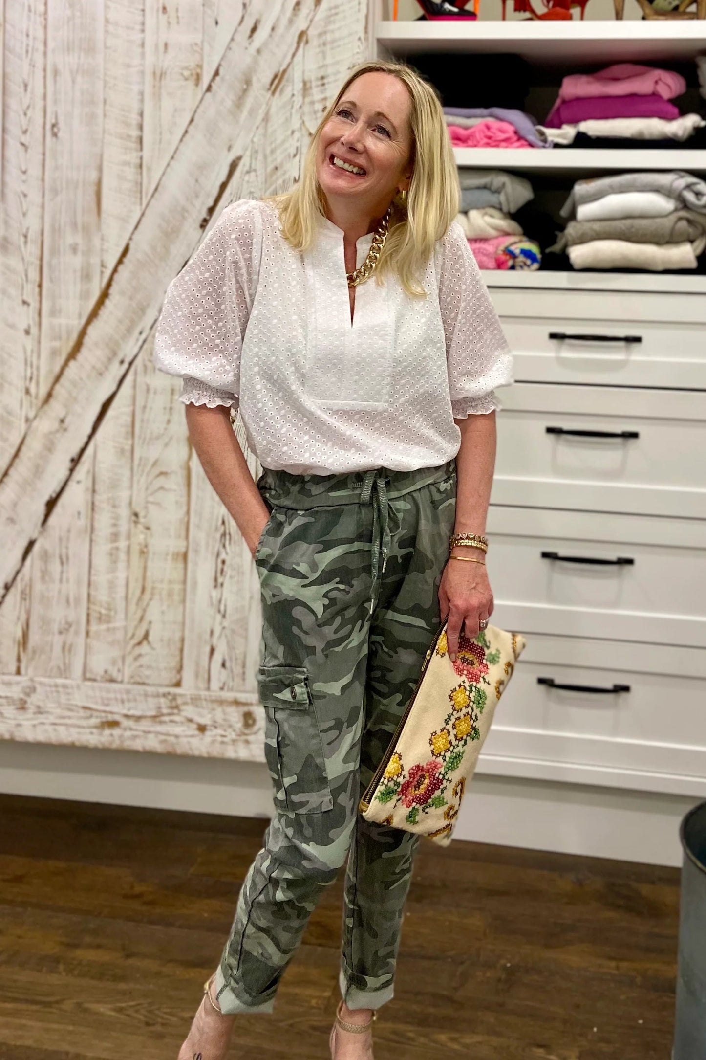 A woman wearing the Eyelet Summer Blouse by Dotty in White, a pullover top with a banded v-neck and 3/4 smock sleeves. She is wearing it with camouflage pants and standing by a closet.