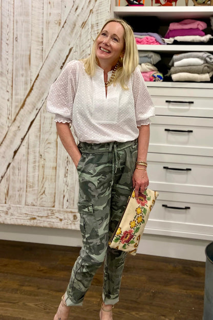 A woman wearing the Eyelet Summer Blouse by Dotty in White, a pullover top with a banded v-neck and 3/4 smock sleeves. She is wearing it with camouflage pants and standing by a closet.