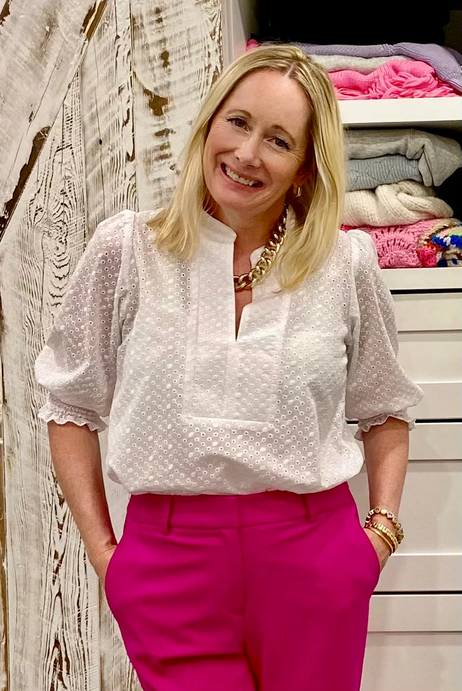 A woman wearing the Eyelet Summer Blouse by Dotty in White, a pullover top with a banded v-neck and 3/4 smock sleeves. She is wearing it with pink pants and standing by a closet. 