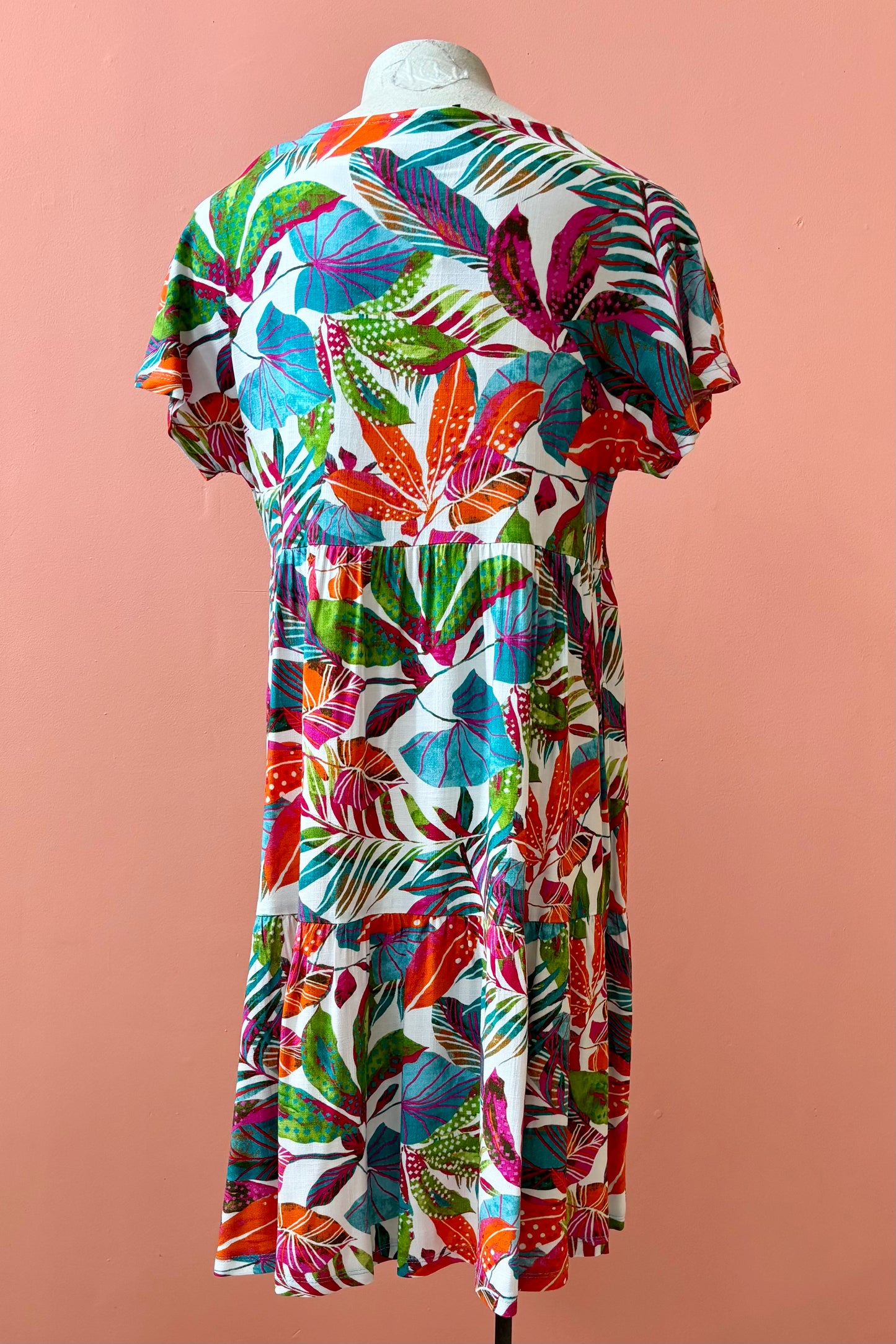 Back view of the Grayson Dress by Pure Essence in Peacock Palms, a knee-length baby doll style dress with a tiered skirt and short sleeves, is shown on a mannequin against a coral  wall. 