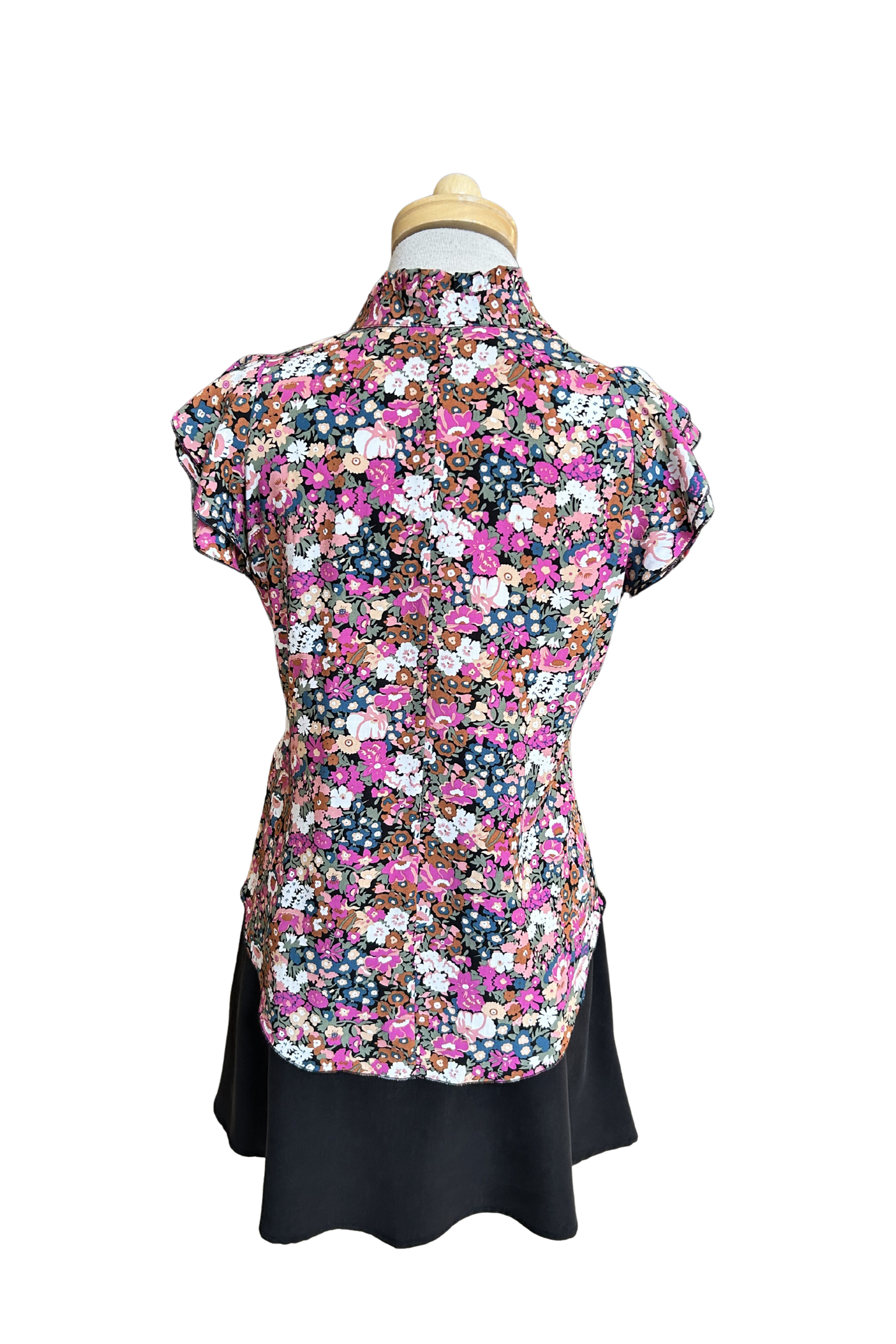 A back view of the Petal Blouse by Dotty in Pink/Blue floral print is shown on a mannequin against a white background. It has short floaty sleeves, a split V-neck, a ruffled collar, and a rounded hi-low hemline. 