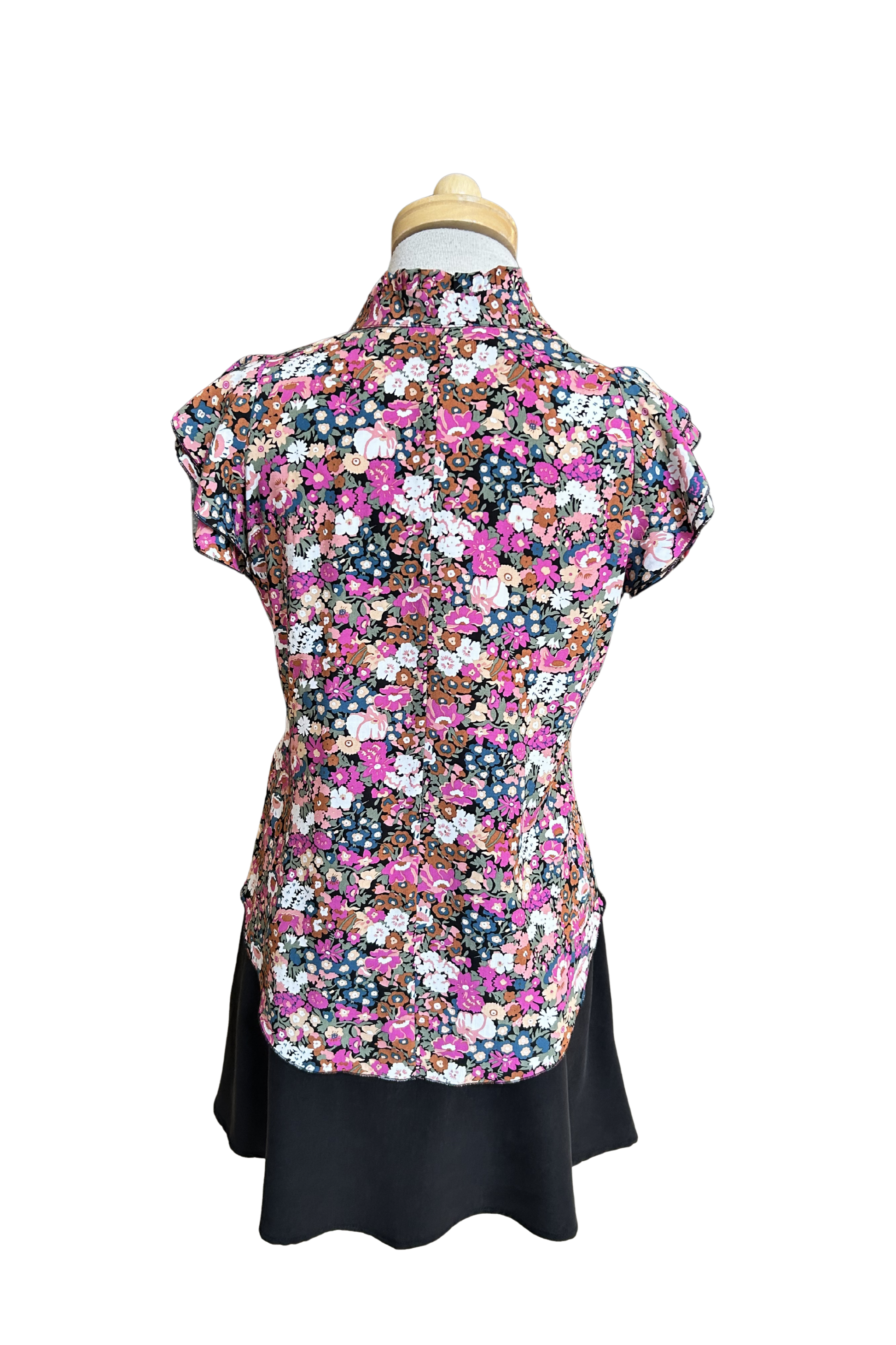 A back view of the Petal Blouse by Dotty in Pink/Blue floral print is shown on a mannequin against a white background. It has short floaty sleeves, a split V-neck, a ruffled collar, and a rounded hi-low hemline. 