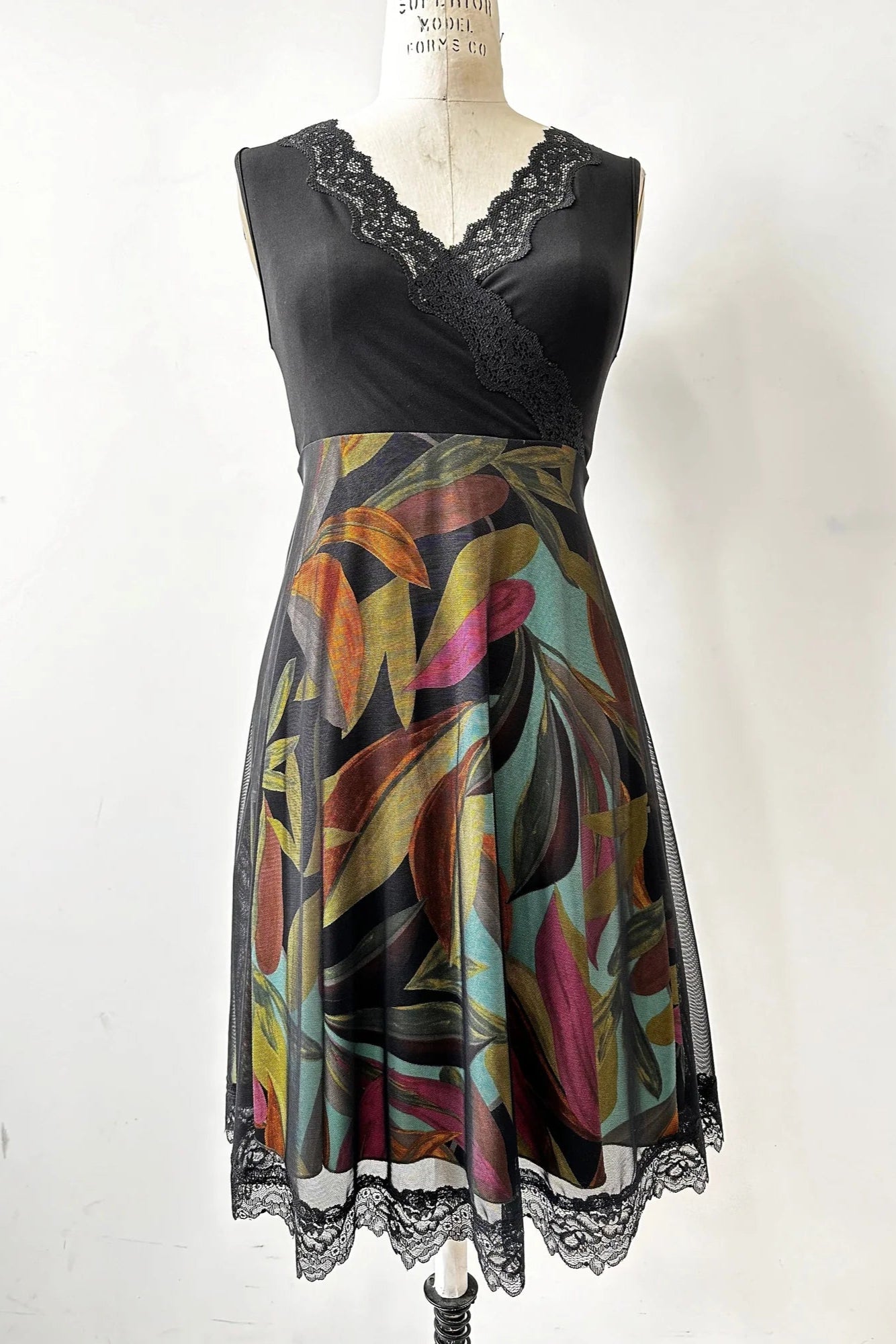 The Deliciosa Dress by Kollontai in Black, a sleeveless dress with an empire waist, lace-trimmed cross-over neckline, and a knee-length tropical print a-line skirt with with a lace-trimmed black mesh overlay, is shown on a mannequin in front of white background 