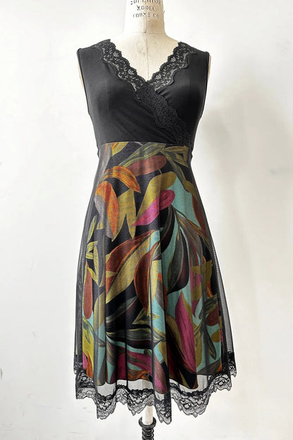 The Deliciosa Dress by Kollontai in Black, a sleeveless dress with an empire waist, lace-trimmed cross-over neckline, and a knee-length tropical print a-line skirt with with a lace-trimmed black mesh overlay, is shown on a mannequin in front of white background 