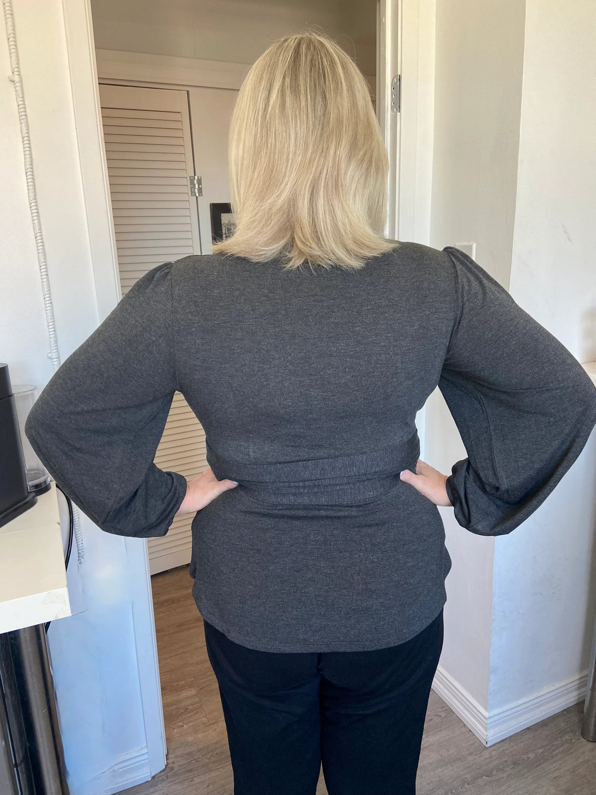 Back view of  woman wearing the Heather Black Bamboo Wrap Top from Dotty, with bracelet length sleeves and adjustable V-neckline. She is wearing it with a black pants and standing in a doorway. 