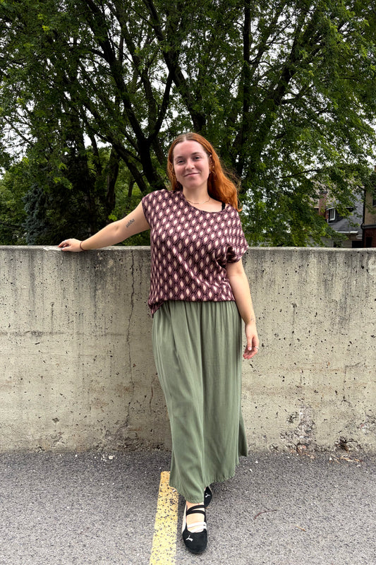 A woman wearing the Fanie Shirt by Kollontai, a relaxed fitting top with cape sleeves and a hi-low hem in a pink and plum geometric print. She is wearing it with green pants and standing outside by a cement wall. 