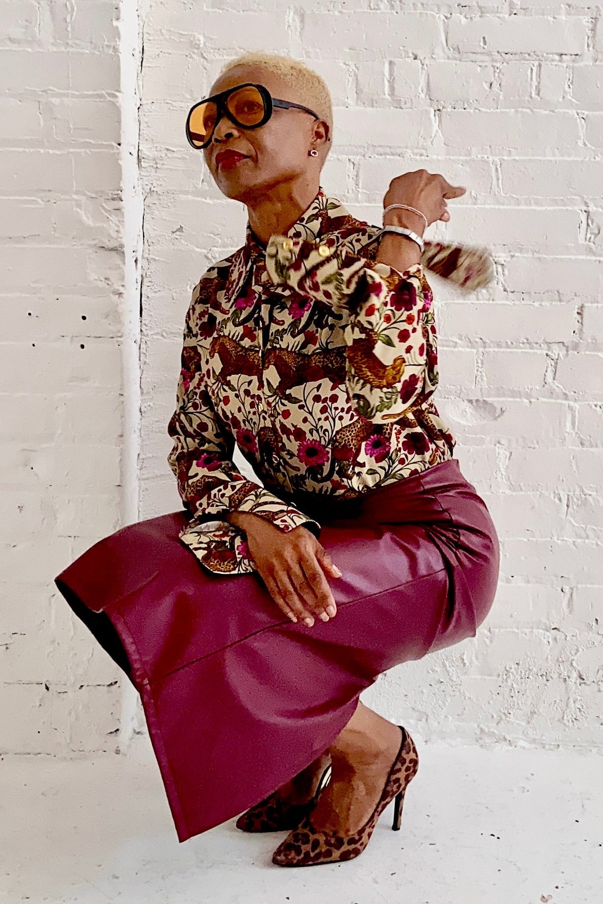 A woman wearing the Untamed Bloom Shirt by Dotty, a menswear-inspired blouse with a floral and animal print, classic button front details, and polished gold buttons. She is wearing it with a pink pleather skirt and crouched down in front of a white brick wall. 