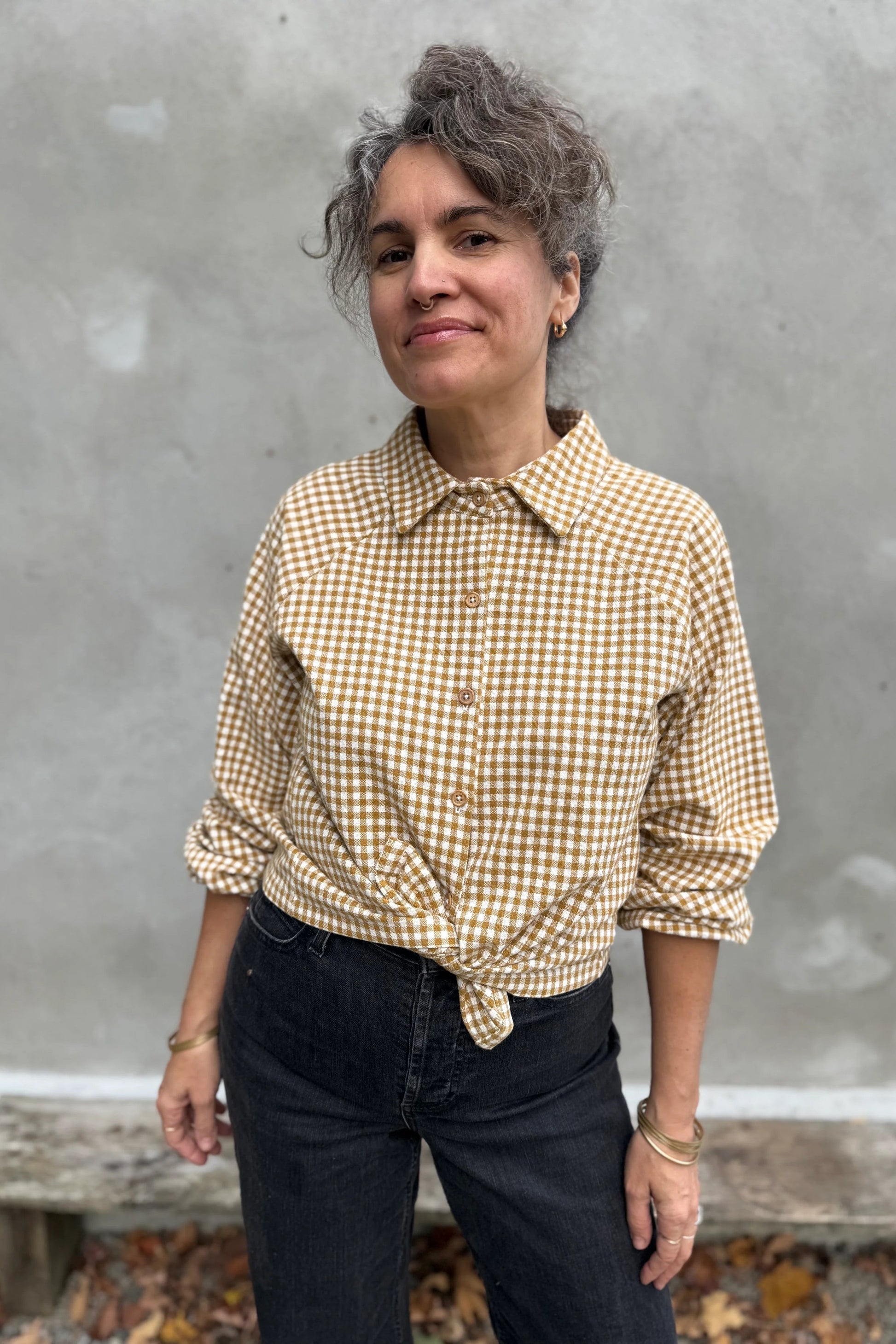 A woman wearing the Acer Blouse by Kazak in Ochre Gingham, a raglan sleeved blouse with a classic collar, button front, and elastic cuffs. She is wearing it with black jeans and standing in front of a grey wall. 