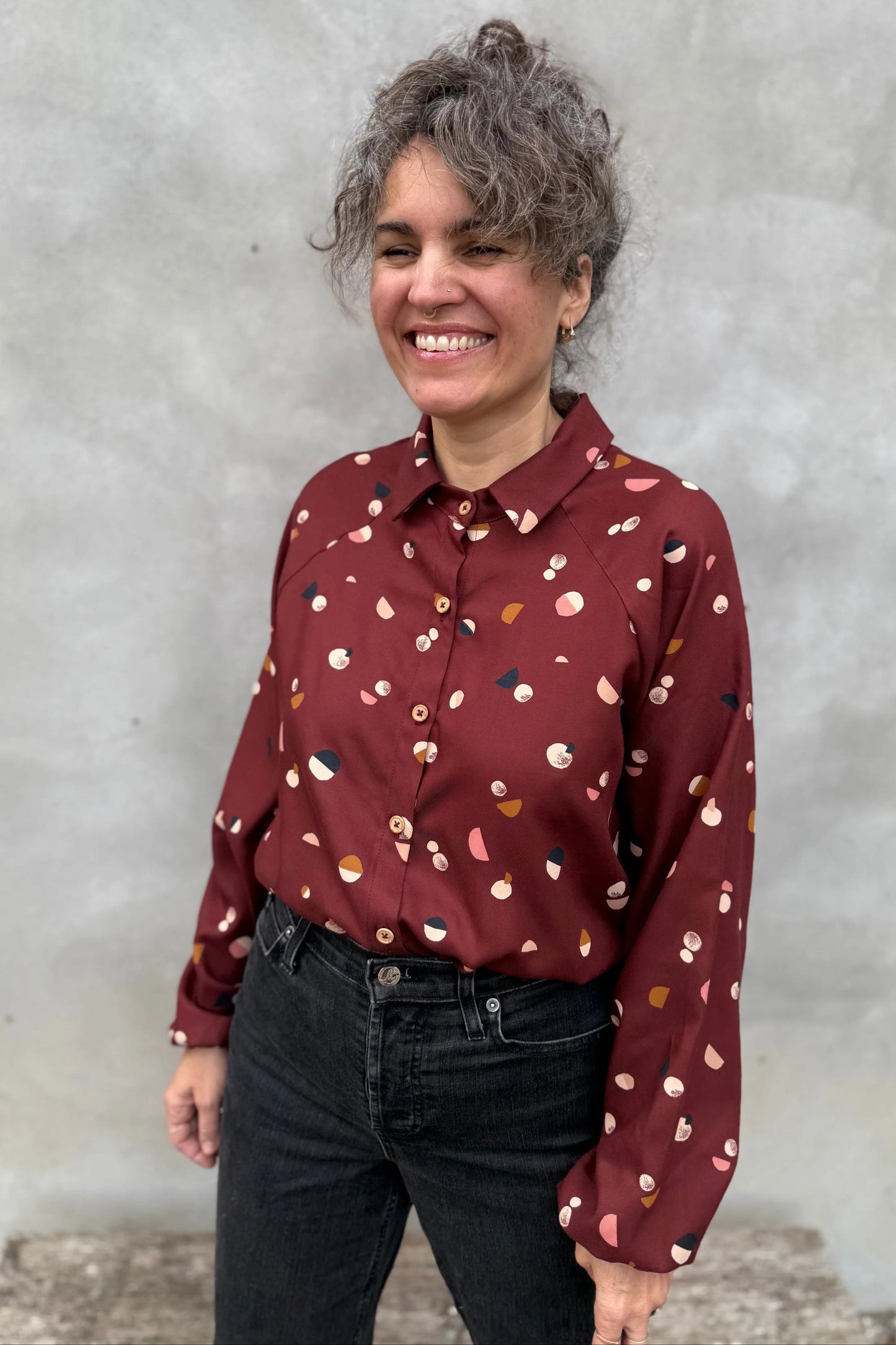 A woman wearing the Acer Blouse by Kazak in Burgundy Moons, a raglan sleeved blouse with a classic collar, button front, and elastic cuffs. She is wearing it with black jeans and standing in front of a grey background. 