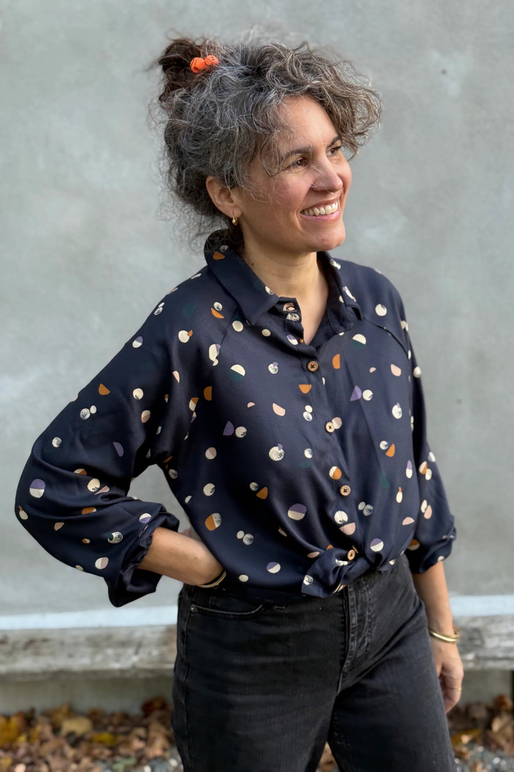 A woman wearing the Acer Blouse by Kazak in Midnight Moons, a raglan sleeved blouse with a classic collar, button front, and elastic cuffs. She is wearing it with black jeans and standing in front of a grey wall. 