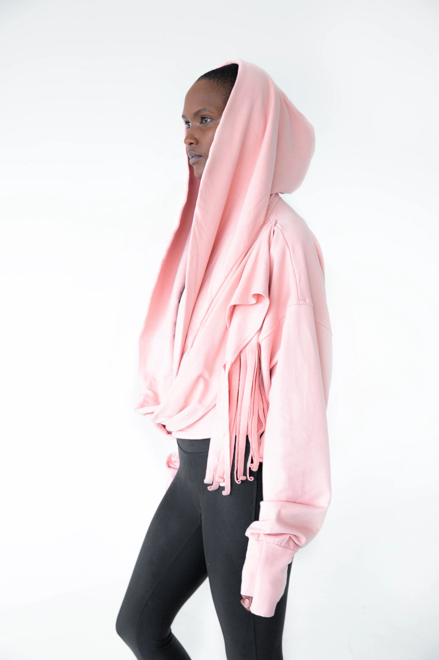 A woman wearing the Heaven Bamboo/Cotton Cropped Wrap from Stacey Martin Lifestyle in Blush Pink, a cropped hoody with a fringed collar scarf that can be tied various ways. She is wearing it with black leggings and standing in a white room. 