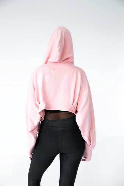 Back view of a woman wearing the Heaven Bamboo/Cotton Cropped Wrap from Stacey Martin Lifestyle in Blush Pink, a cropped hoody with a fringed collar scarf that can be tied various ways. She is wearing it with black leggings and standing in a white room. 