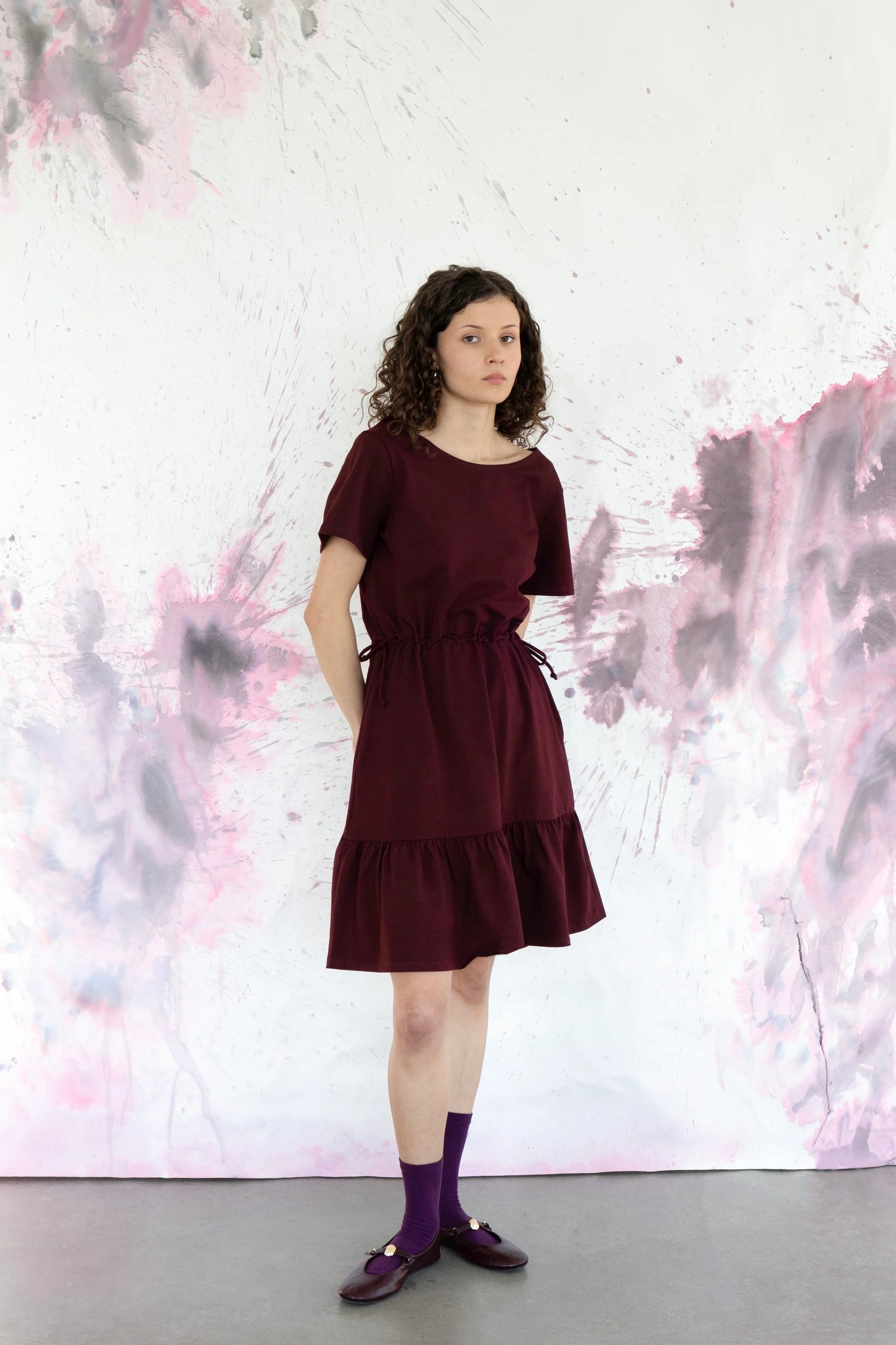 A woman wearing the Épervière Dress by Eve Lavoie in Burgundy, a reversible dress with short sleeves, a V-neck on one side and a round neck on the other, a flounced hem, and ties at the sides of the waists. She is standing against an abstract purple and grey wall. 
