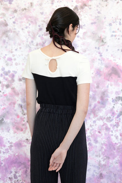 Back view of a woman wearing the Gentiane Top by Eve Lavoie in Black/White, a two-toned fitted tee with a sweetheart-effect neckline on one side and a cutout and tie detail on the other. She is wearing it with pinstripe pants and standing in front of a purple and grey abstract background. 
