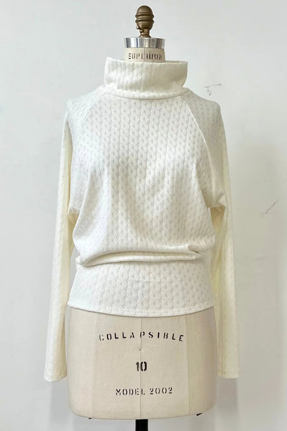 The Plume Sweater by Kollontai in White, a cable knit turtleneck with long tapered sleeves and a narrow hem for a bloused look, is shown on a mannequin in front of a white background. 