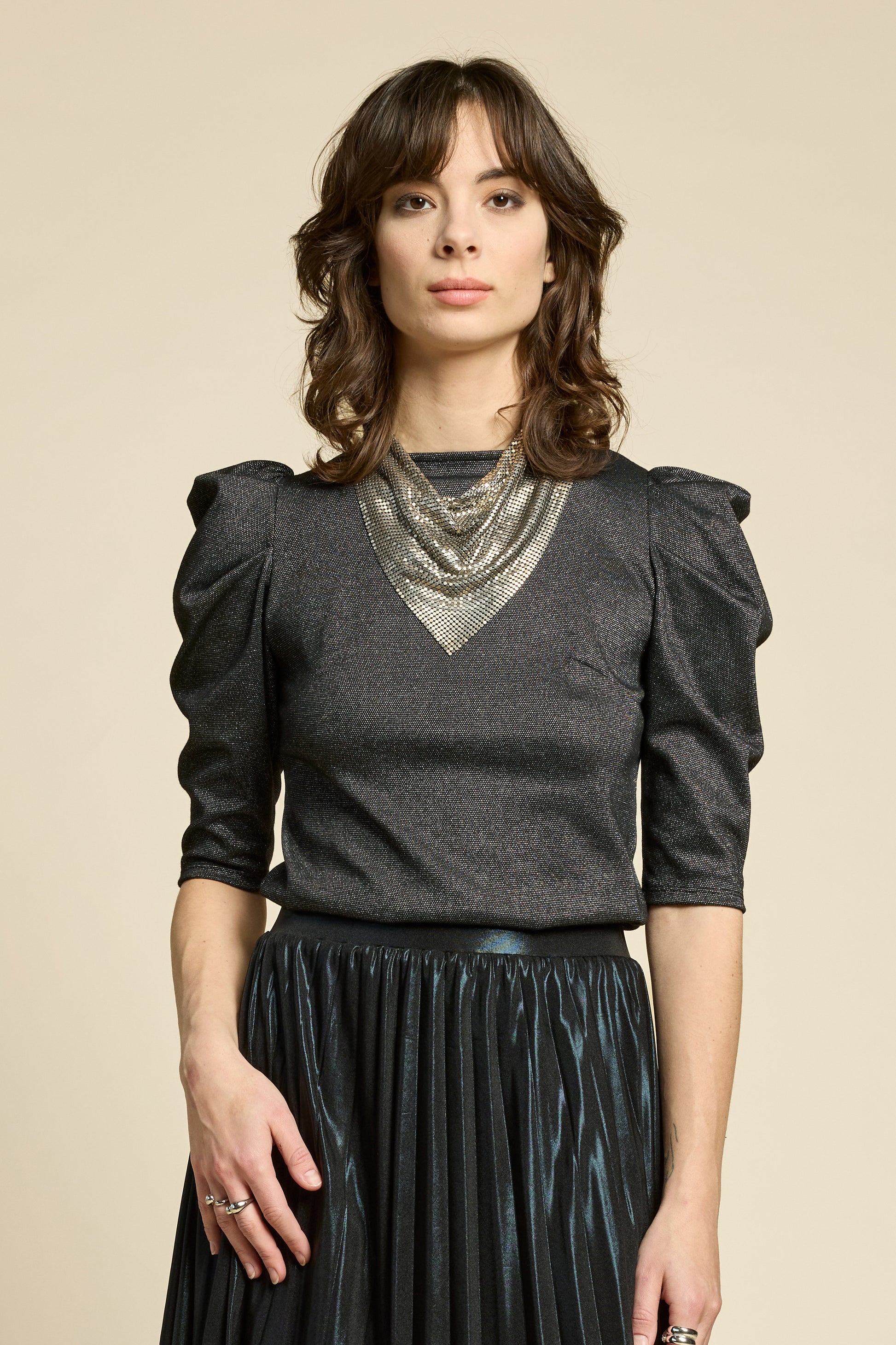A woman wearing the Insomnie Top by Cokluch in Facet Black, a metallic piece with 3/4 puffed sleeves, darts a the bust, and a slim fit. She is wearing it with the Feyer Skirt in Glow Black Knit and standing in front of a beige background. 