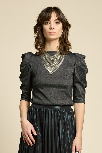 A woman wearing the Insomnie Top by Cokluch in Facet Black, a metallic piece with 3/4 puffed sleeves, darts a the bust, and a slim fit. She is wearing it with the Feyer Skirt in Glow Black Knit and standing in front of a beige background. 