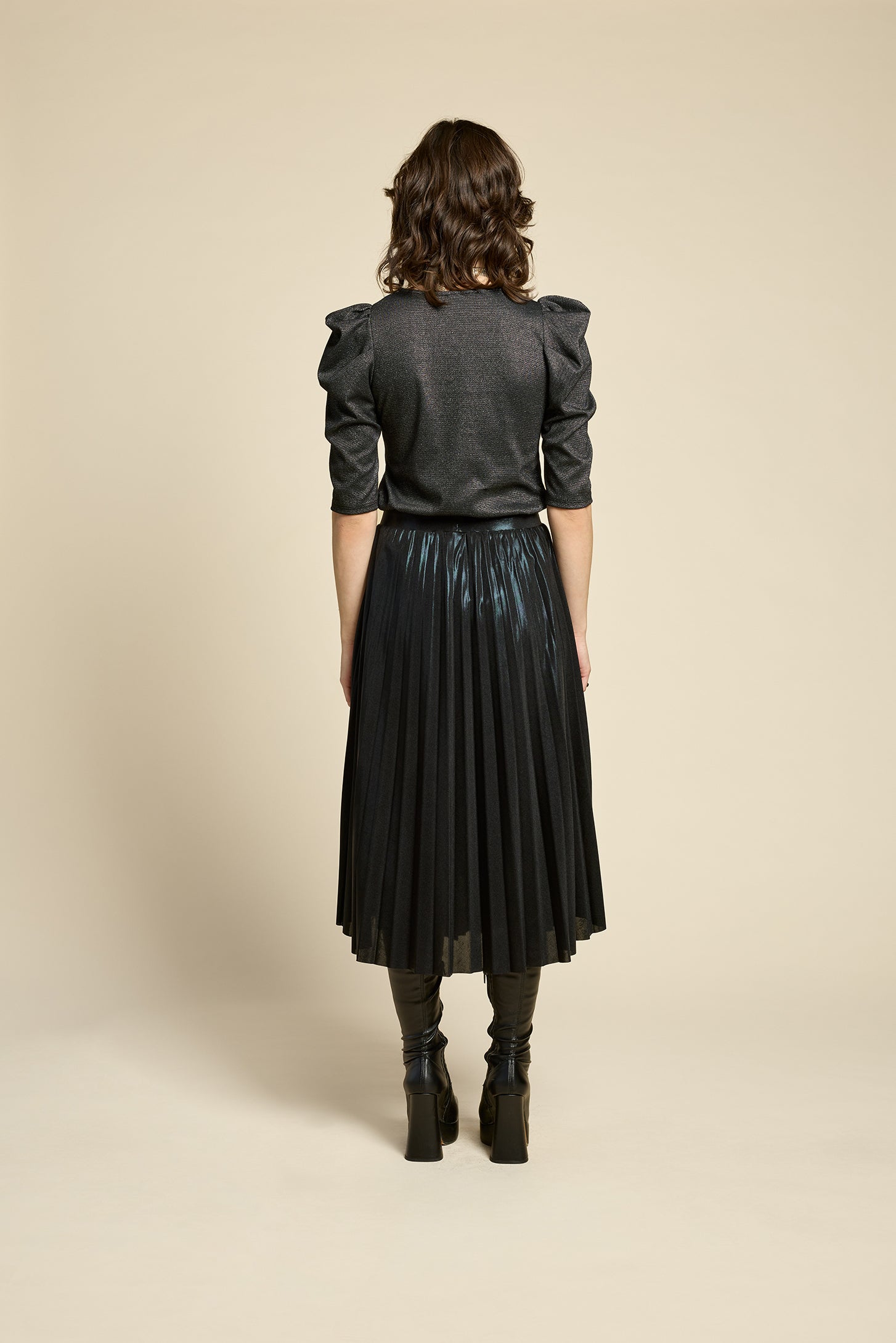 Back view of a woman wearing the Insomnie Top by Cokluch in Facet Black, a metallic piece with 3/4 puffed sleeves, darts at the bust, and a slim fit. She is wearing it with the Feyer Skirt in Glow Black Knit and standing in front of a beige background. 