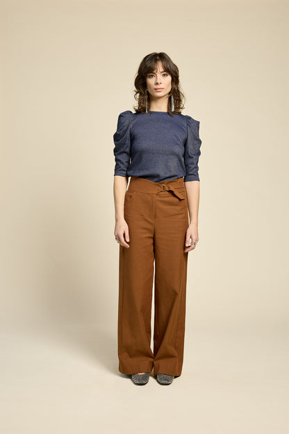 A woman wearing the Insomnie Top by Cokluch in Facet Twilight, a metallic piece with 3/4 puffed sleeves, darts at the bust, and a slim fit. She is wearing it with brown pants and standing in front of a beige background. 