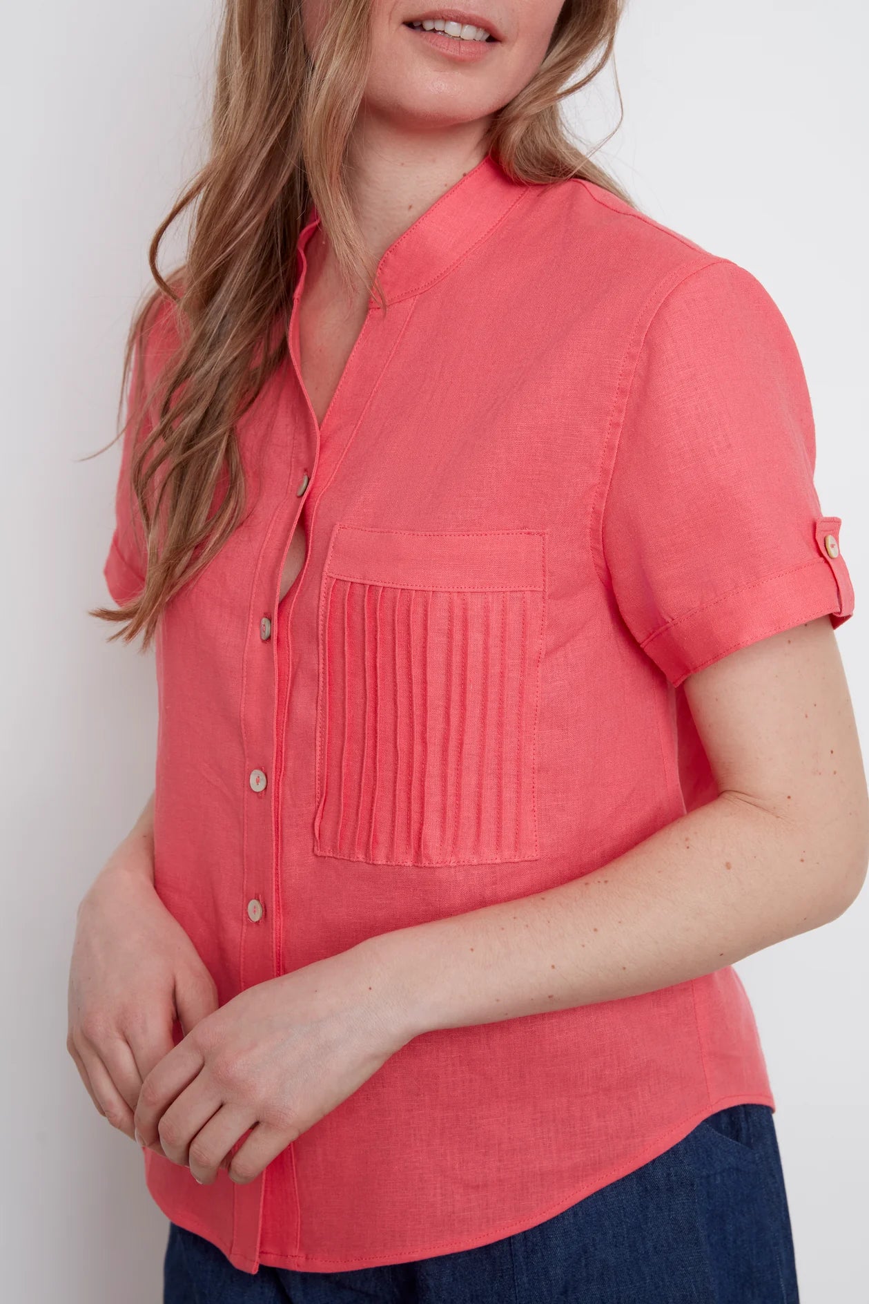 Close up view of a woman wearing the Jasmine Short Sleeve Linen Shirt by Julei in Coral, with a mandarin collar, pleated pocket, and cuff and tab sleeves. She is wearing it with jeans and standing in front of a white background. 