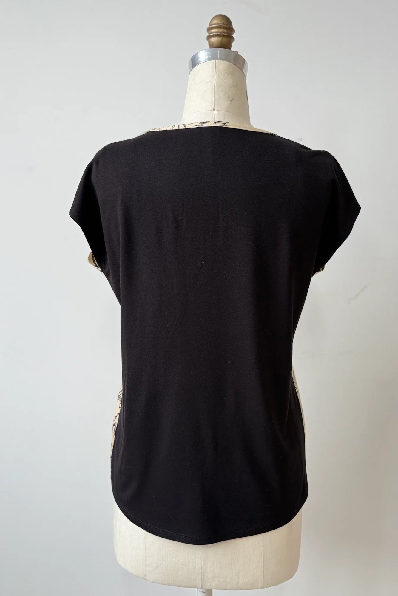 A back view of the Arty T-Shirt by Kollontai in Cats/Beige print, with a round neck, short sleeves, and a slight hi-low hemline, and a solid black back, is shown on a mannequin against a grey background. 