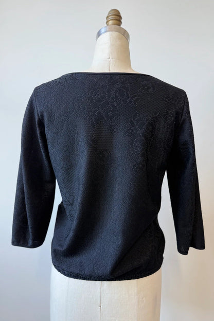 A back view of the Celestine Blouse By Kollontai in Black, featuring black satin overlaid with black lace, is shown on a mannequin in front of a white background. It has a cropped silhouette with an elastic waist, and 3/4 sleeves. 