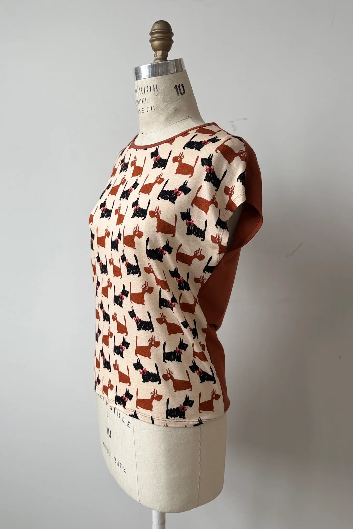 A side view of the Arty T-Shirt by Kollontai in Dogs/Rust print, with a round neck, short extended sleeves, and a slight hi-low hemline, and a solid rust back, is shown on a mannequin against a grey background. 