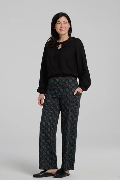 A woman wearing the Elma Pants by Kollontai in Turquoise on black print, straight legged pants with a wide elastic waist and large slant pockets. She is wearing them with a black top and standing in front of a grey background. 