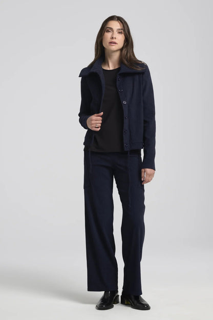 A woman wearing the Hermione Pants by Kollontai in Navy, stretchy corduroy pants with a wide pull-one waistband, wide legs, and angled patch pockets. She is wearing them with a dark top and jacket and standing in front of a grey background. 