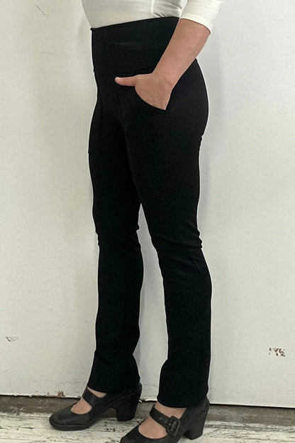 Side waist down view of a woman wearing the Bari Pants by Kollontai, black pants with a slim fit, wide pull-on waistband, and front patch pockets, in front of a white wall. 