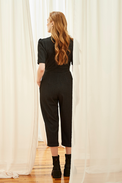 Back view of a woman wearing the Lexie Jumpsuit by Melow in Black, featuring short sleeves, a V-neck, invisible front zipper, elastic waist, and 34/ length legs in a diagonal stripe fabric. She is standing in front of a white curtain. 